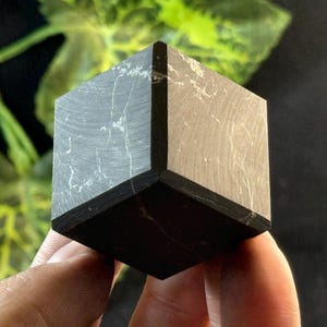 Könnte beinhalten: Ein dunkelgrauer, würfelförmiger Stein mit weißen Adern. Der Würfel wird in einer Hand gehalten, mit einem verschwommenen grünen und schwarzen Hintergrund. Der Stein hat ein poliertes Aussehen und ist wahrscheinlich ein Dekorationsartikel oder ein Mineralexemplar.