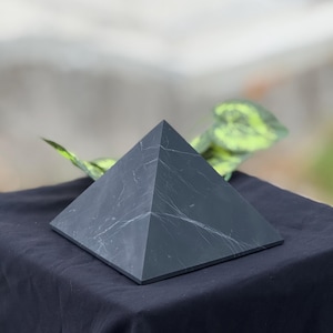 以下が含まれることがあります： 白い縞模様のある濃い灰色のピラミッドが黒い表面に置かれています。ピラミッドは正方形の底面と4つの三角形の側面を持っています。背景は緑の葉でぼやけています。