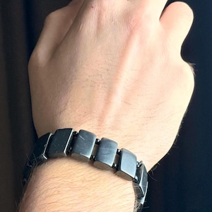 May include: A dark gray bracelet composed of rectangular, connected links. The bracelet is worn on a wrist, showcasing its geometric design and metallic appearance. The bracelet's links are approximately 0.75 inches in length.