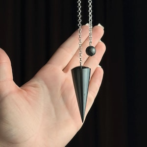 Può includere: Un pendolo nero a forma conica e una piccola sfera, sospeso da una catena d'argento. Il pendolo è tenuto su uno sfondo scuro.