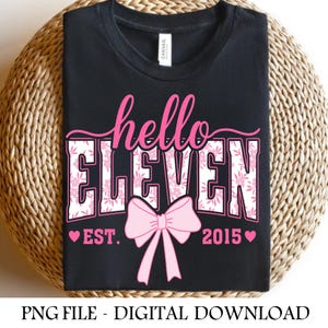 May include: Black t-shirt with the text "hello ELEVEN" in pink script and floral print. A pink bow is centered below the text, with "EST. 2015" and hearts. The shirt is folded on a woven surface.