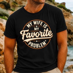 May include: A black short-sleeved t-shirt featuring a circular graphic design with the text "MY WIFE IS MY FAVORITE PROBLEM" in white and light orange lettering, accented by small stars and horizontal lines within the circular border.