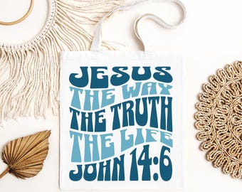 May include: White tote bag featuring the text "JESUS THE WAY THE TRUTH THE LIFE JOHN 14:6" in shades of blue. The bag has two white handles and is made of a soft fabric. The design is centered on the bag.