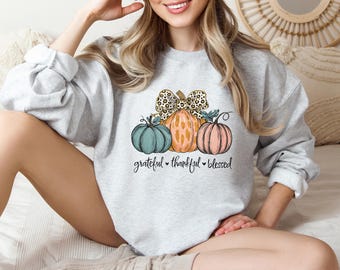 Könnte beinhalten: Hellgraues Sweatshirt mit einer Grafik von drei Kürbissen in Türkis, Orange und Rosa, mit einer Schleife im Leopardenmuster. Darunter stehen die Worte "grateful, thankful, blessed".