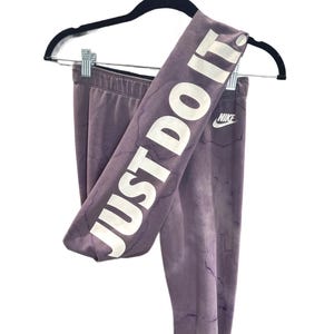 Pode incluir: Leggings roxos com o texto "JUST DO IT" em letras brancas grandes. O logotipo da Nike é visível. As leggings estão penduradas em um cabide preto em um fundo branco.