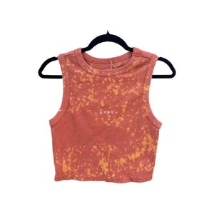 May include: A coral-colored, ribbed crop top with a bleached, orange pattern. The sleeveless top has a scoop neck and the word "ROXY" printed in white. The top is hanging on a black hanger.