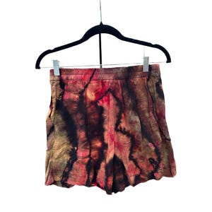 May include: Tie-dye shorts in red, brown, and black hang from a black hanger. The shorts have a pleated design and an elasticated waistband. The fabric appears soft and lightweight, suitable for warm weather.