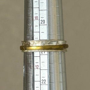 May include: A silver and gold ring is placed on a ring sizer. The ring has a textured silver band and a gold band. The ring sizer has measurements in inches and is used to determine the ring size.