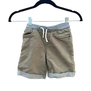 May include: Khaki shorts with a grey elasticated waistband and drawstring. The shorts have two front pockets and rolled-up cuffs. The garment is hanging on a black hanger against a white background.