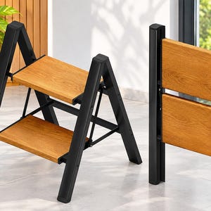 May include: A two-step folding ladder featuring a black metal frame and two light brown wood-grain steps. The ladder is shown in both an open, functional position and a closed, upright position for storage, highlighting its collapsible design.
