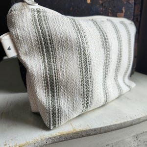 May include: A rectangular, off-white cosmetic bag with vertical stripes in a muted green hue. The bag is made of a textured fabric and features a zipper closure with a small, white tag attached to the side. The bag is approximately 8 inches wide.