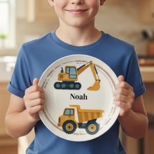 May include: A white plate featuring yellow construction vehicles, an excavator and a dump truck, with the name "Noah" printed in black. The plate has a brown decorative border.