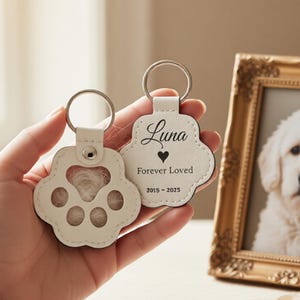 May include: Two white leather paw-shaped keychains with silver key rings. One keychain has a paw print cutout filled with fur. The other keychain has the name "Luna", a heart, and "Forever Loved 2015-2025" printed on it. A framed photo is in the background.