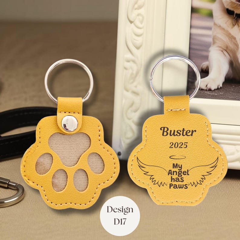 May include: Yellow paw-shaped keychain with a silver keyring. One keychain has a clear window, the other has the text "Buster 2025 My Angel has Paws" and wings. The design code "D17" is also visible.