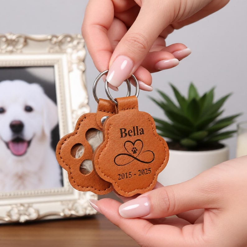 May include: Two brown leather paw-shaped keychains with silver rings. One keychain has a paw print cutout, the other has the name "Bella", a heart with a paw print, and the years "2015-2025" engraved. A framed dog portrait is in the background.