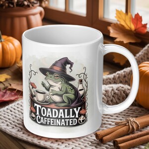 May include: White ceramic mug featuring a green frog wearing a witch hat, holding a coffee cup. The mug has the text "Toadally Caffeinated" and is decorated with mushrooms and a potion bottle. Perfect for coffee or tea.