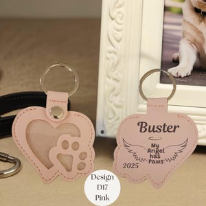 May include: Pink heart-shaped keychain with a paw print and a photo window. The keychain on the right has the name "Buster", angel wings, and the text "My Angel has Paws 2025". The design is labeled "Design D17 Pink".