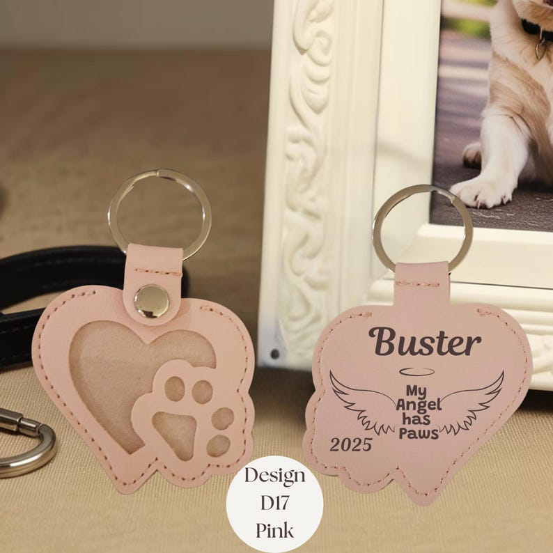 May include: Pink heart-shaped keychain with a paw print and a photo window. The keychain on the right has the name "Buster", angel wings, and the text "My Angel has Paws 2025". The design is labeled "Design D17 Pink".