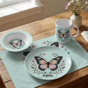 May include: A light blue ceramic bowl, plate, and mug set with a butterfly design and the name "Sophia". The butterfly has black and pink wings, surrounded by a floral wreath. The set is on a matching placemat.