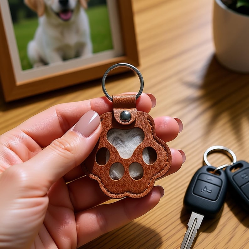 May include: A brown leather paw print keychain with a silver key ring. The keychain has a heart-shaped cutout filled with white fur. A framed photo of a dog and car keys are in the background.