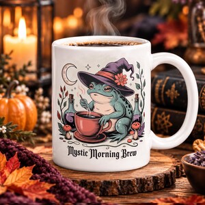 May include: A white ceramic mug featuring a graphic of a green frog wearing a purple witch hat, holding a red teacup. The illustration includes potion bottles, mushrooms, a crescent moon, and the text Mystic Morning Brew in black lettering.