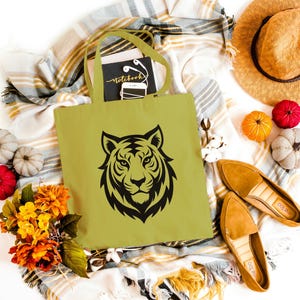 May include: A light green tote bag with a black tiger face design. The bag is surrounded by fall-themed decorations, including pumpkins, flowers, a straw hat, and tan loafers. A notebook with the word "Notebook" is inside the bag.