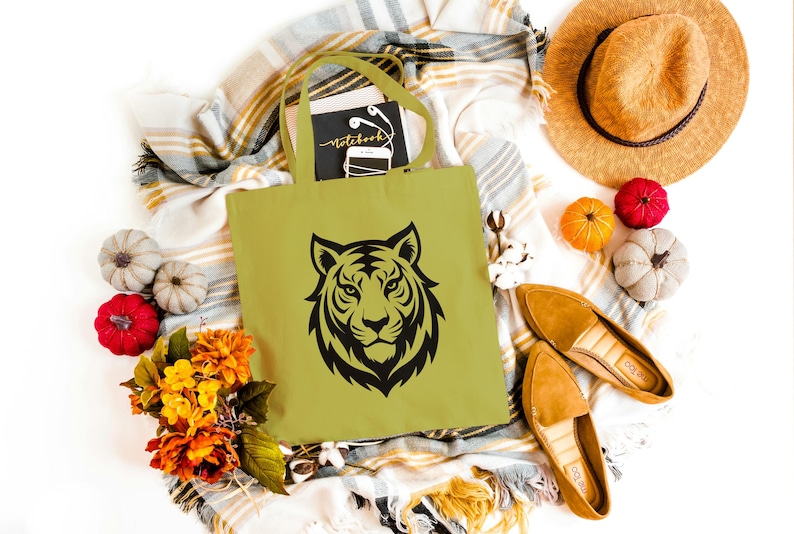 May include: A light green tote bag with a black tiger face design. The bag is surrounded by fall-themed decorations, including pumpkins, flowers, a straw hat, and tan loafers. A notebook with the word "Notebook" is inside the bag.