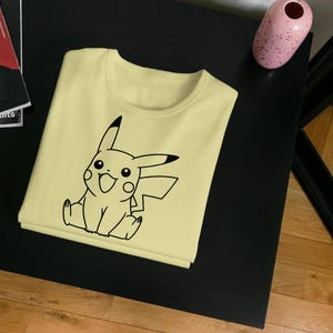 May include: A light yellow t-shirt featuring a black outline of Pikachu, a popular character. The shirt is folded neatly on a black surface, with a magazine and a pink vase in the background. The shirt is a casual wear item.