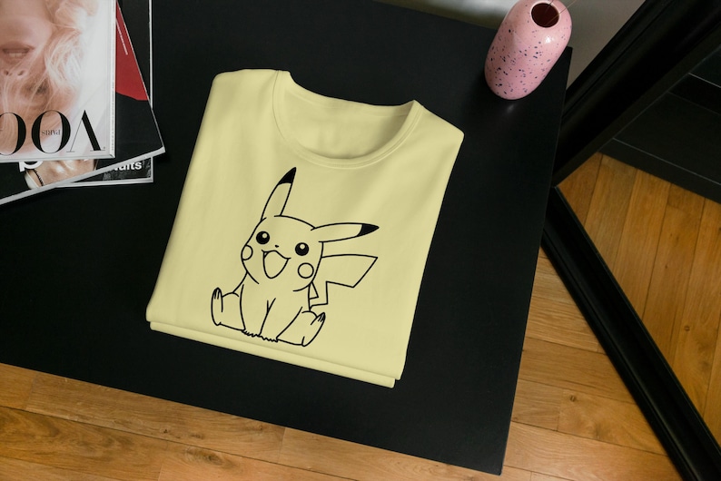 May include: A light yellow t-shirt featuring a black outline of Pikachu, a popular character. The shirt is folded neatly on a black surface, with a magazine and a pink vase in the background. The shirt is a casual wear item.