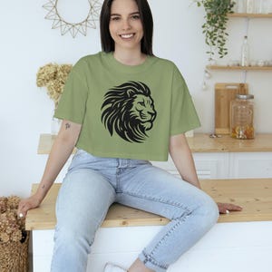 May include: Sage green crop top featuring a black lion head design. The shirt is a casual style, suitable for everyday wear. The model is wearing light wash jeans and white sneakers.