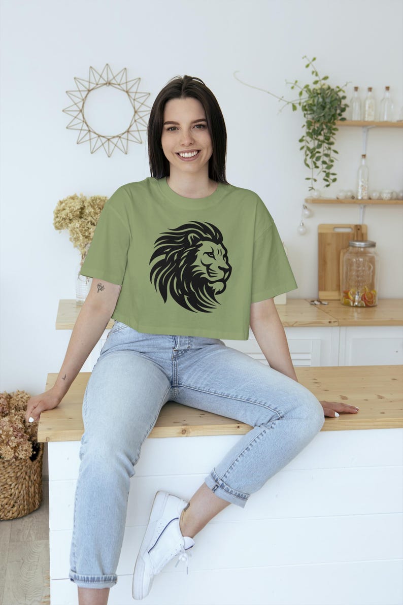 May include: Sage green crop top featuring a black lion head design. The shirt is a casual style, suitable for everyday wear. The model is wearing light wash jeans and white sneakers.