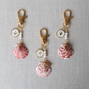 Pode incluir: Três chaveiros com fechos dourados, cada um com um pingente de concha. As conchas são rosa e brancas, com detalhes dourados e pequenas pérolas. Um chaveiro tem um pingente de cruz, outro uma bússola e o terceiro uma letra.