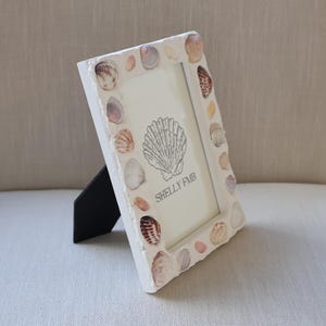 May include: White picture frame decorated with various seashells. The frame has a black stand and features a seashell illustration with the text "SHELLY FMB". The frame is a decorative item for coastal or beach-themed decor.
