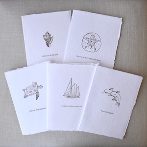 Peut inclure: Cinq cartes de vœux blanches aux bords déchiquetés, chacune avec un dessin au trait noir différent de la vie marine. Les dessins comprennent un coquillage, un dollar des sables, une tortue, un voilier et des dauphins. Chaque carte a un message basé sur un jeu de mots.