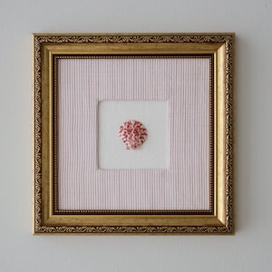 Könnte beinhalten: Ein gerahmtes Kunstwerk mit einem goldfarbenen, verzierten Rahmen. Im Inneren befindet sich ein quadratisches weißes Stoffstück, das auf einem rosa-weiß gestreiften Hintergrund zentriert ist. Ein kleines, rot-weißes Stickdesign befindet sich in der Mitte des weißen Quadrats.