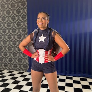 May include: A woman in a Captain America-themed costume. The outfit features a navy blue top with a white star, matching shorts, a red and white corset, and red wrist cuffs. She is smiling, with a shield on her back.