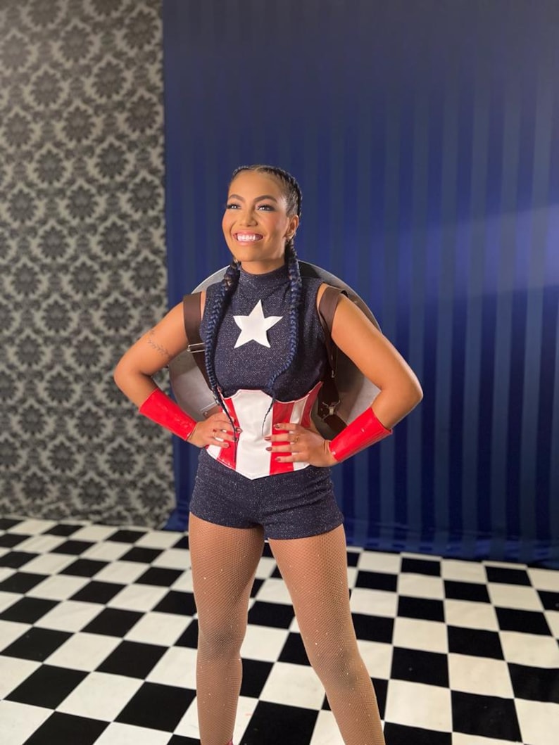 May include: A woman in a Captain America-themed costume. The outfit features a navy blue top with a white star, matching shorts, a red and white corset, and red wrist cuffs. She is smiling, with a shield on her back.