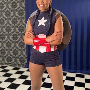 May include: A navy blue superhero costume with a white star, red and white belt, and red wrist cuffs. The costume includes a shield and matching shorts. The background has a blue and white checkered floor.