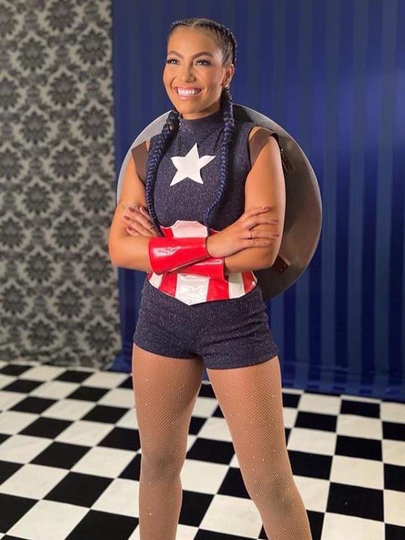 May include: A navy blue superhero costume with a white star, red and white belt, and red wrist cuffs. The costume includes a shield and matching shorts. The background has a blue and white checkered floor.