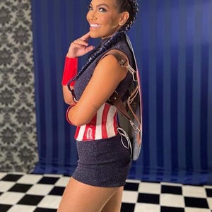 May include: A woman in a navy sequined mini-dress and a red, white, and blue top. She has a black and brown leather accessory on her back. Her hair is styled in braids. She is smiling and looking to the side.