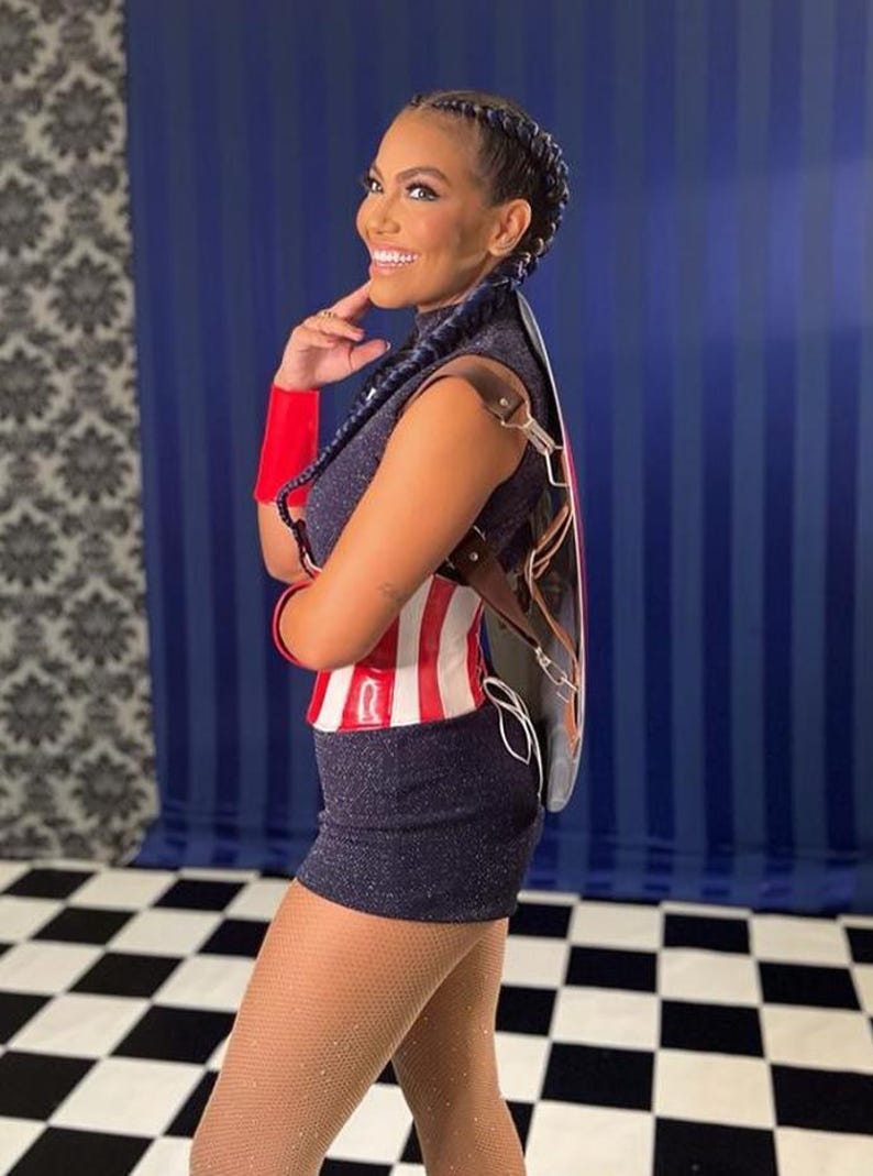 May include: A woman in a navy sequined mini-dress and a red, white, and blue top. She has a black and brown leather accessory on her back. Her hair is styled in braids. She is smiling and looking to the side.