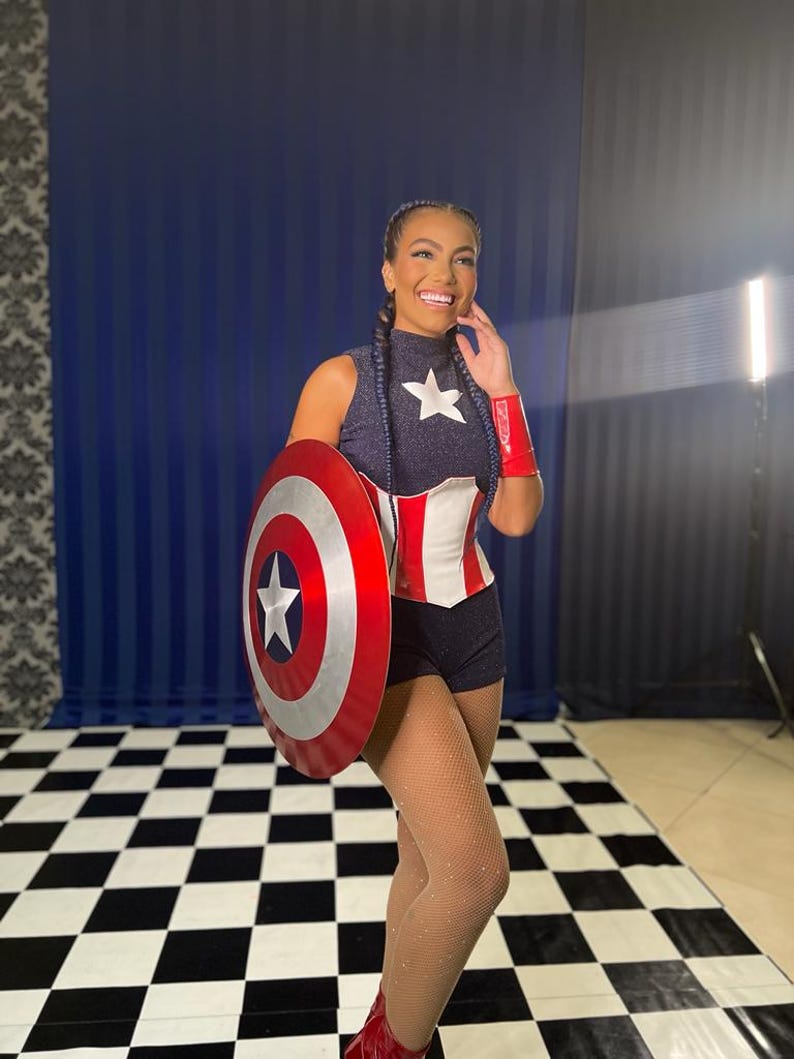 May include: A person in a Captain America-themed costume, holding a shield. The outfit features a navy top with a white star, a red and white corset, and black shorts. Red wristbands and boots, plus patterned tights complete the look.