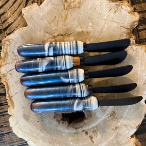 May include: Five butter knives with black blades and handles featuring a blue, white, and black marbled design. The knives are arranged on a light-colored, organic-shaped wooden surface.