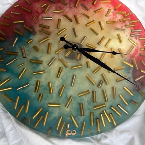 May include: A round wall clock with a colorful resin face. The face is decorated with brass bullet casings and features black clock hands. The clock face has a gradient of colors, including red, blue, and yellow.