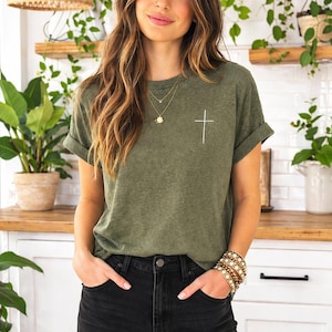 May include: Olive green t-shirt with a white cross graphic. The shirt has rolled-up sleeves and is paired with black distressed jeans. A gold necklace and bracelet are also visible.