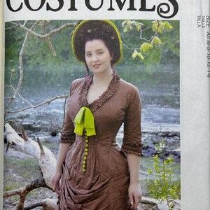 May include: McCall's Costumes pattern cover featuring a woman in a brown dress with a green bow and hat. The dress has a ruffled neckline and sleeves, with a row of green buttons down the front. The background shows a natural outdoor scene.