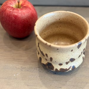 May include: A handmade ceramic mug with a speckled beige glaze and dark brown spots. A red apple with a green stem sits next to the mug on a wooden surface. The mug is a small, round shape.