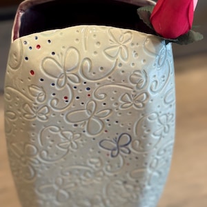 May include: A light-colored ceramic vase with an embossed butterfly pattern. The vase has a dark purple interior and is decorated with red and blue dots. A red rose is placed on the rim of the vase.