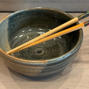May include: A handmade ceramic bowl in shades of blue and green, with a textured exterior and a smooth interior. Two wooden chopsticks with decorative ends rest across the bowl. The bowl is designed for serving food.