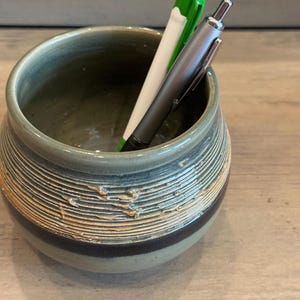 May include: A handmade ceramic pen holder with a textured, ridged design in shades of green, brown, and tan. Three pens, one green, one white, and one silver, are inside the holder. The pen holder is a decorative desk accessory.